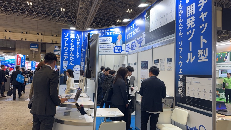 Japan IT Week の BlueLamp の展示の場。「チャット型開発ツール」の看板の前で来場者が画面を見ている